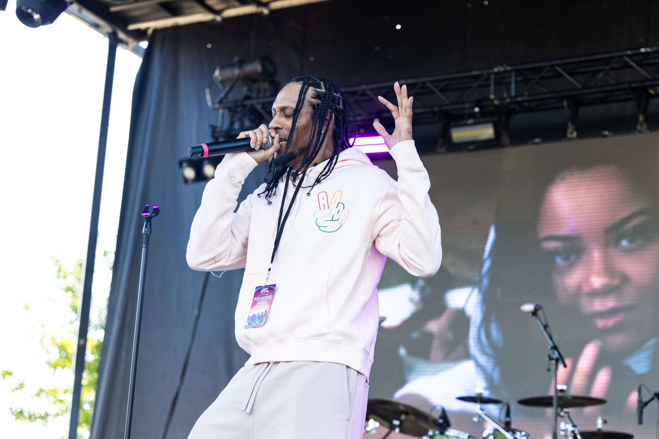 Person with long braids singing into a microphone on stage, wearing a white hoodie, with a large screen showing a woman's face behind them.