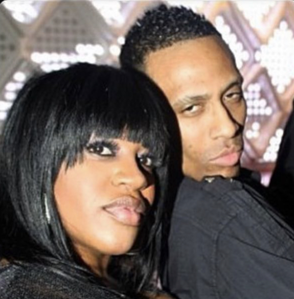 Two people pose closely, one with black hair and bangs, the other with short curly hair, in a patterned background.