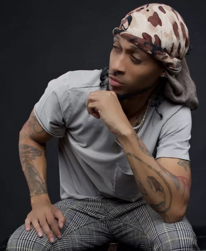Young man with a patterned headscarf, gray t-shirt, and plaid pants, sitting with hand near face, against dark background.