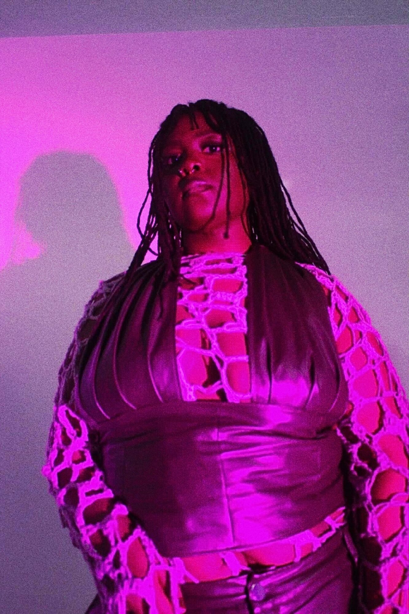 Person with long braided hair wearing a purple top with cut-out sleeves, standing against a purple background.