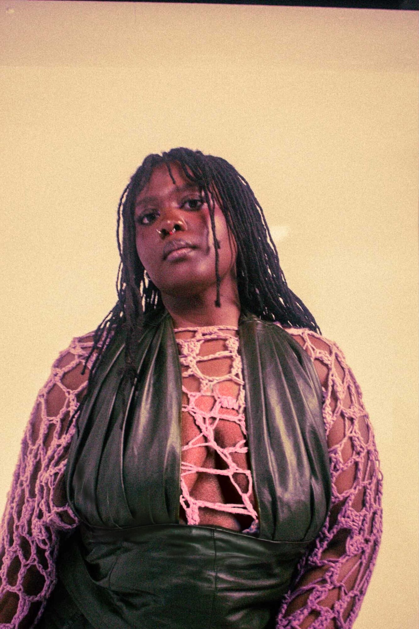 Woman with long braided hair wearing a pink lace top and black leather vest against a plain background.