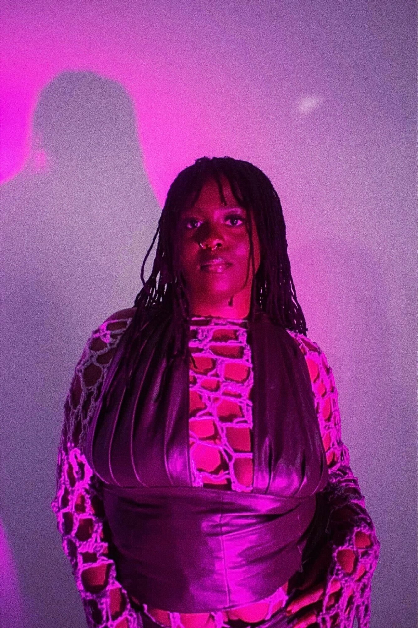 Woman with long dreadlocks wearing a patterned top, illuminated by purple light, standing against a purple background.