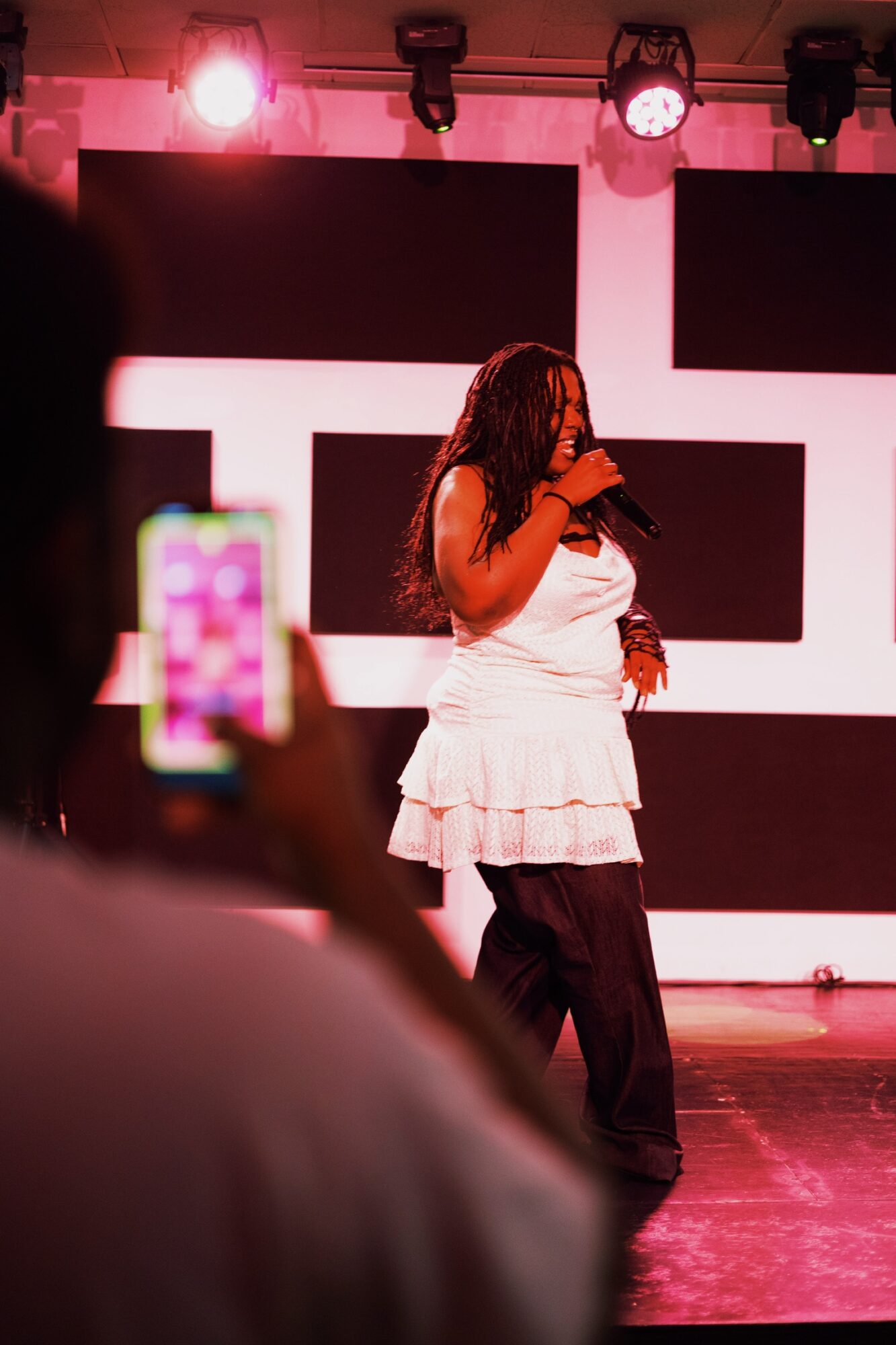 Woman with long hair speaking into a microphone on stage with pink lighting and geometric background.