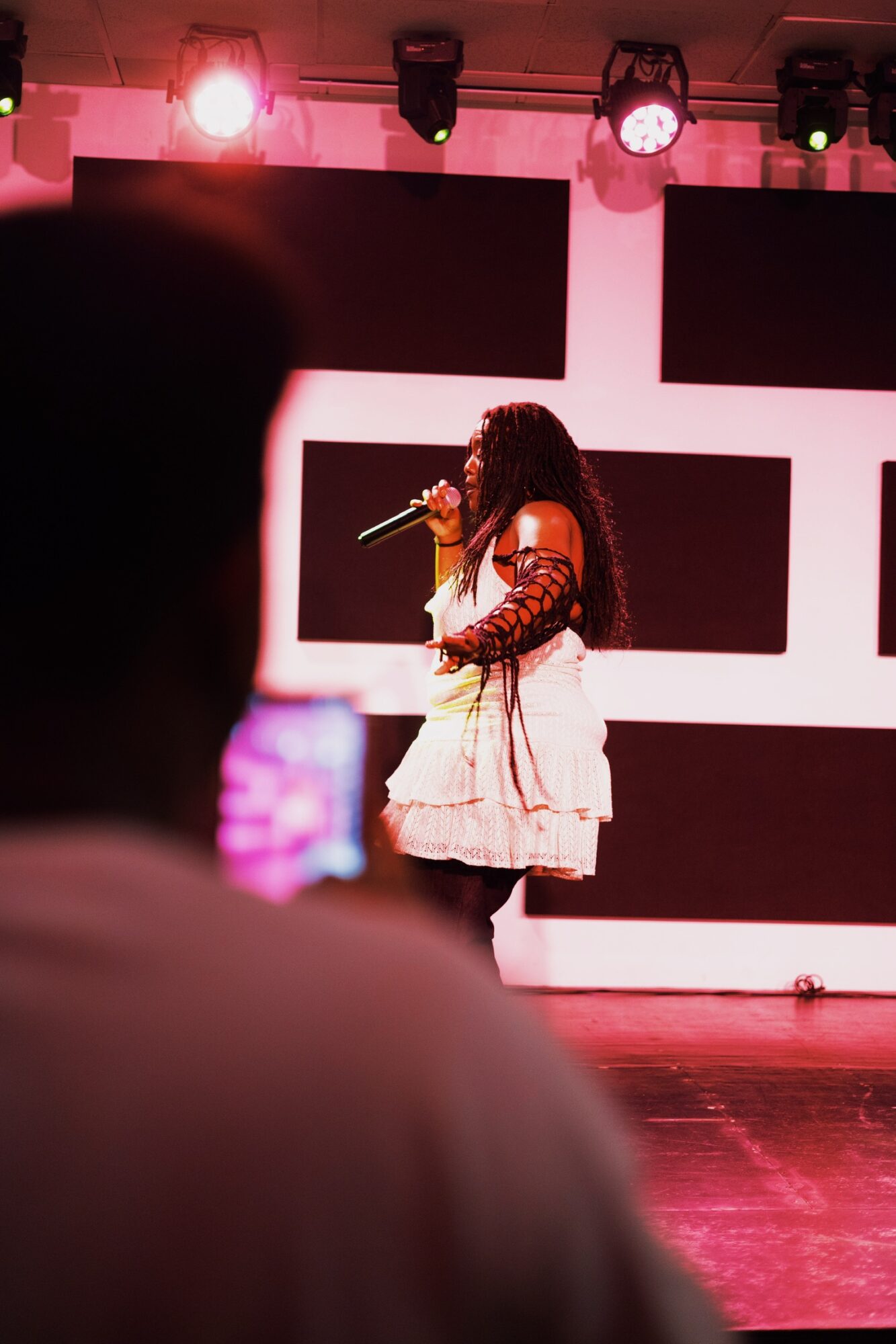 Performer with curly hair singing into a microphone on stage, audience in foreground, stage lights overhead.