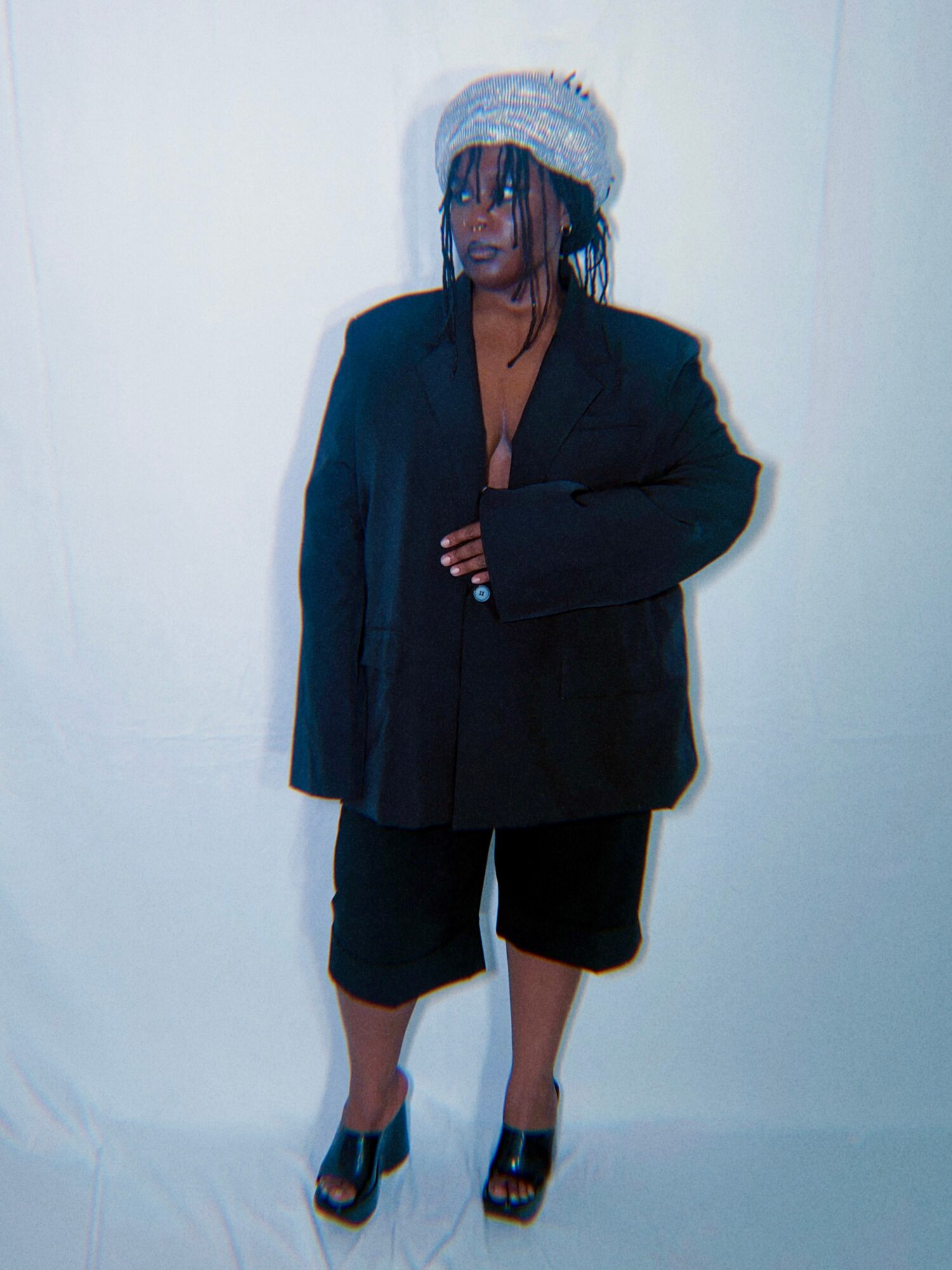 Person wearing a hat, oversized black blazer, black shorts, and high-heeled sandals, standing against a plain white background.