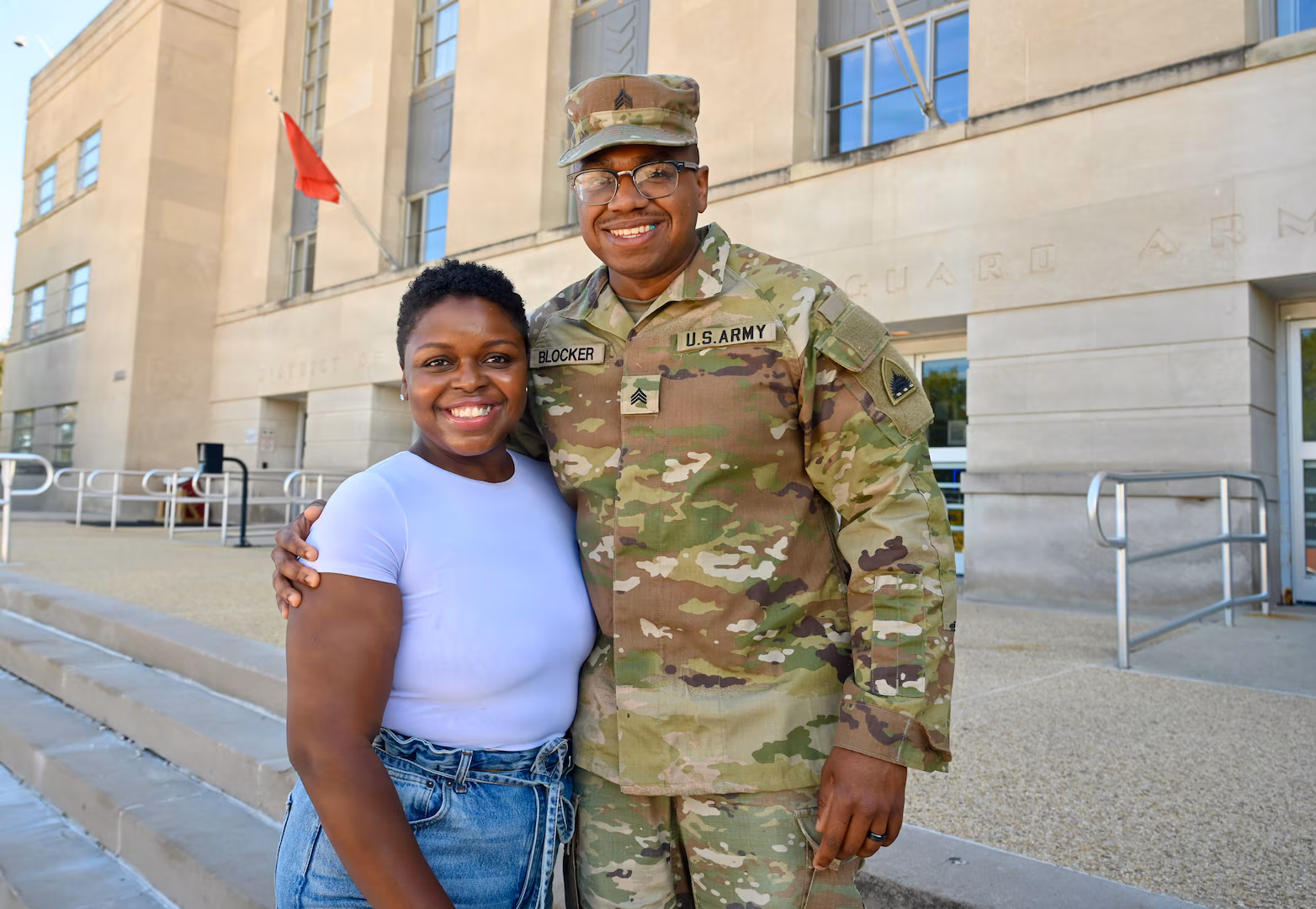 A woman and a soldier in uniform stand outside a building, smiling and posing together.