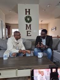 Two men sit on a sofa in a living room, with a large vertical sign reading 'HOME' behind them, and a person taking a photo.