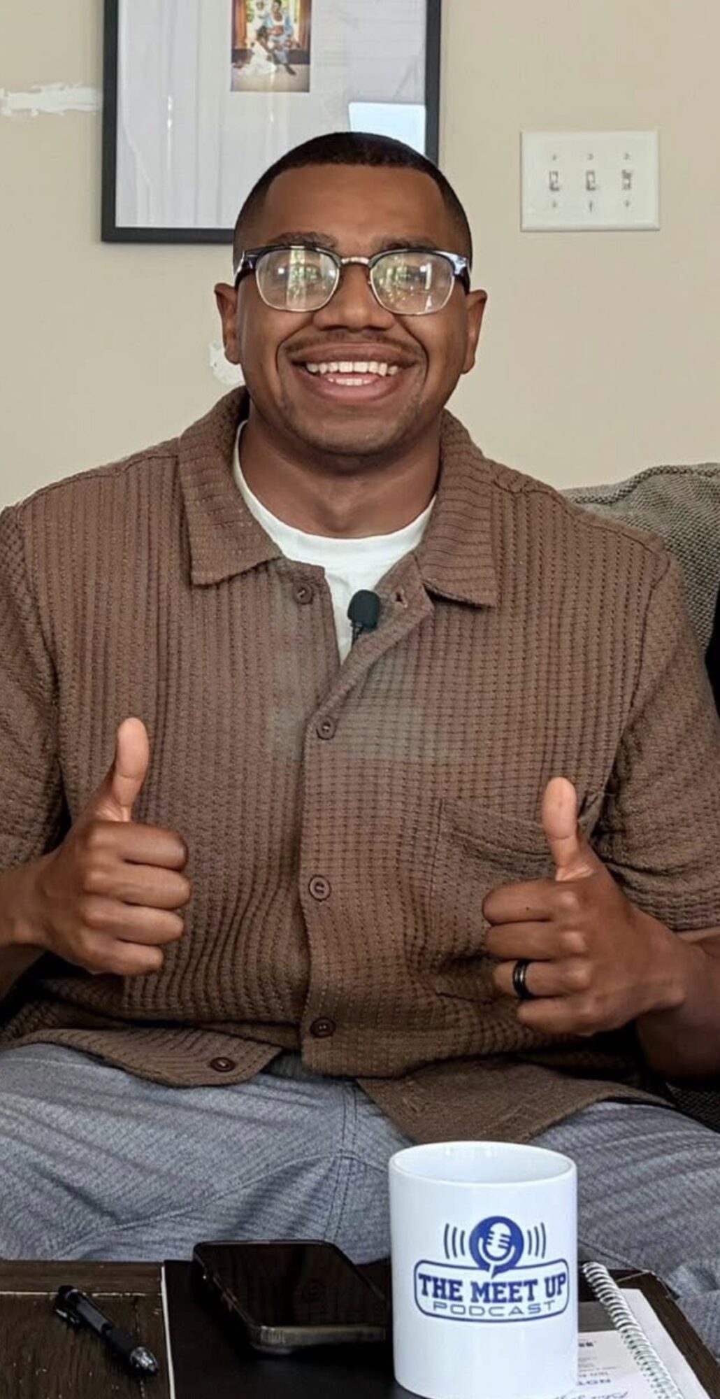 Smiling man wearing glasses and a brown shirt giving two thumbs up, sitting indoors with a mug and phone in front.