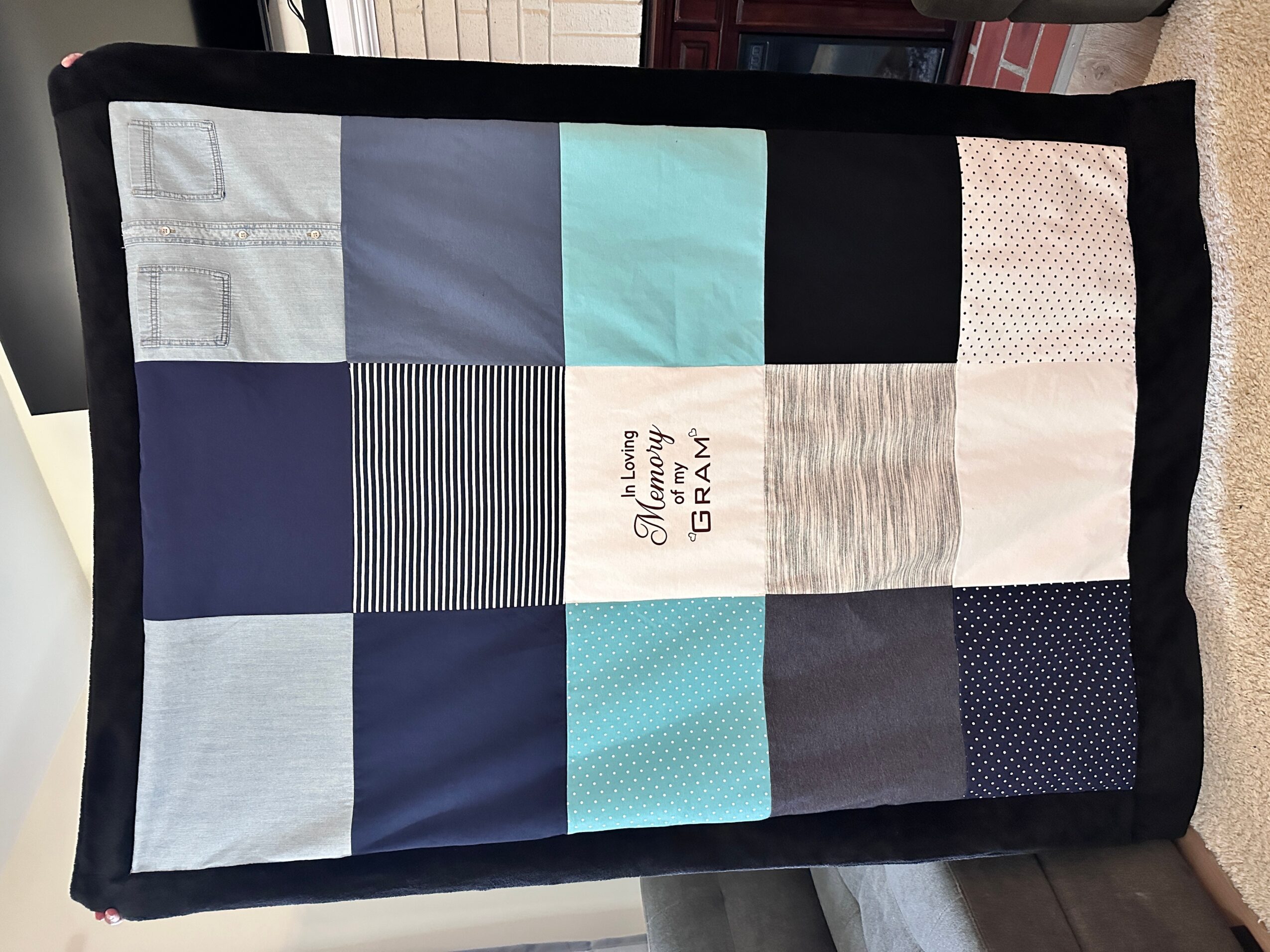 A quilt with various blue, black, and beige fabric patches, some with patterns, and a central patch with text.