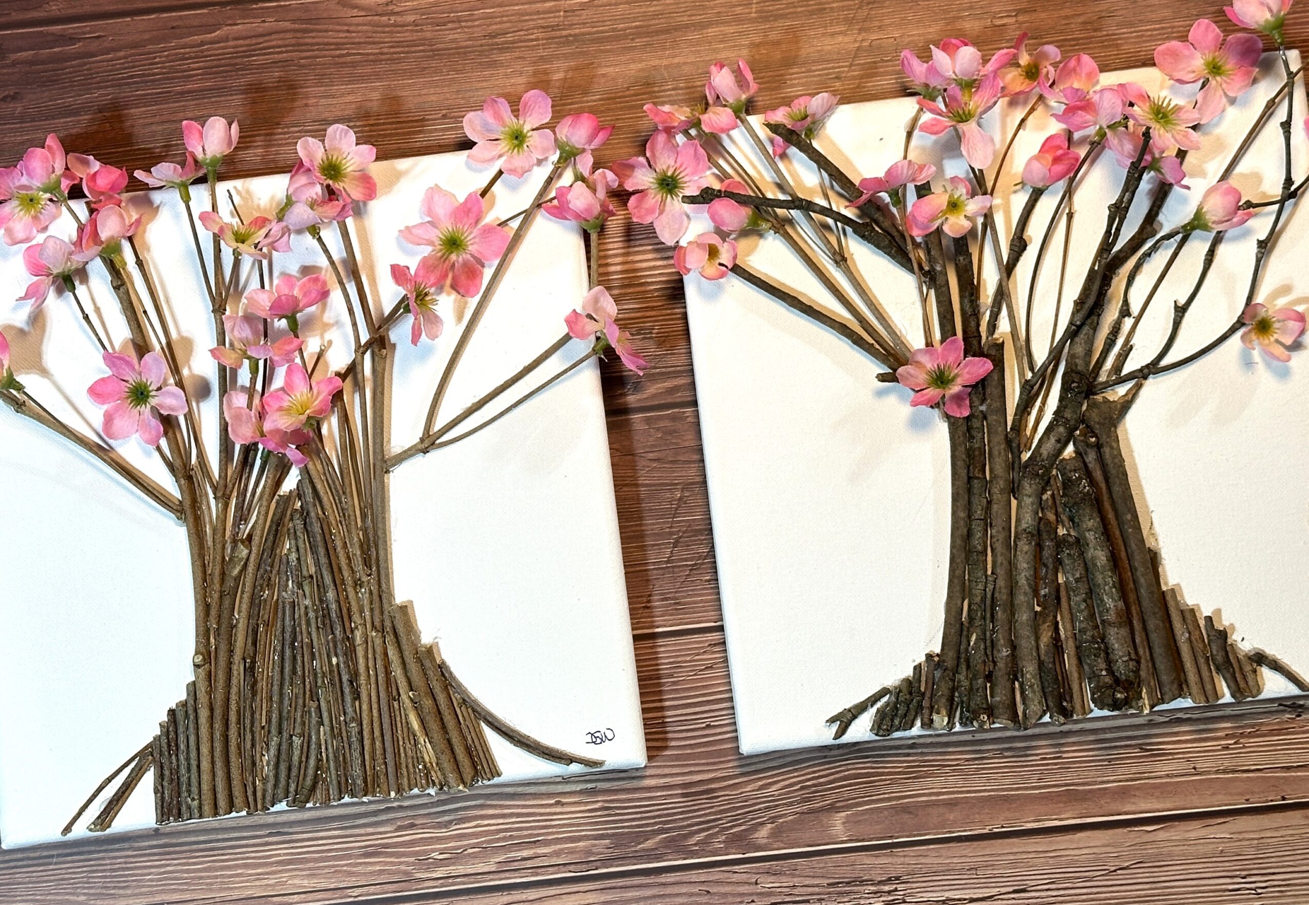 Two paper crafts of trees with pink flowers and brown trunks on white backgrounds, placed on a wooden surface.