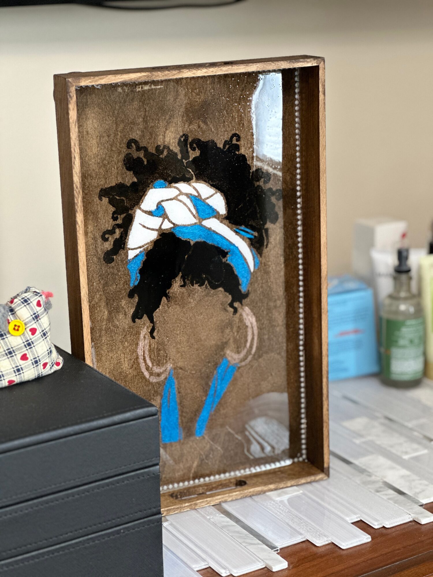 Framed artwork of a woman with natural hair, headwrap, and earrings, displayed on a table with various items nearby.