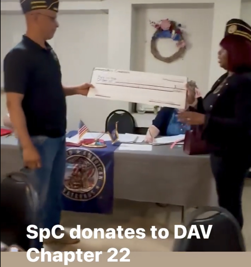 Person in uniform hands large check to woman in uniform at table with American flag and seal, wreath on wall behind.