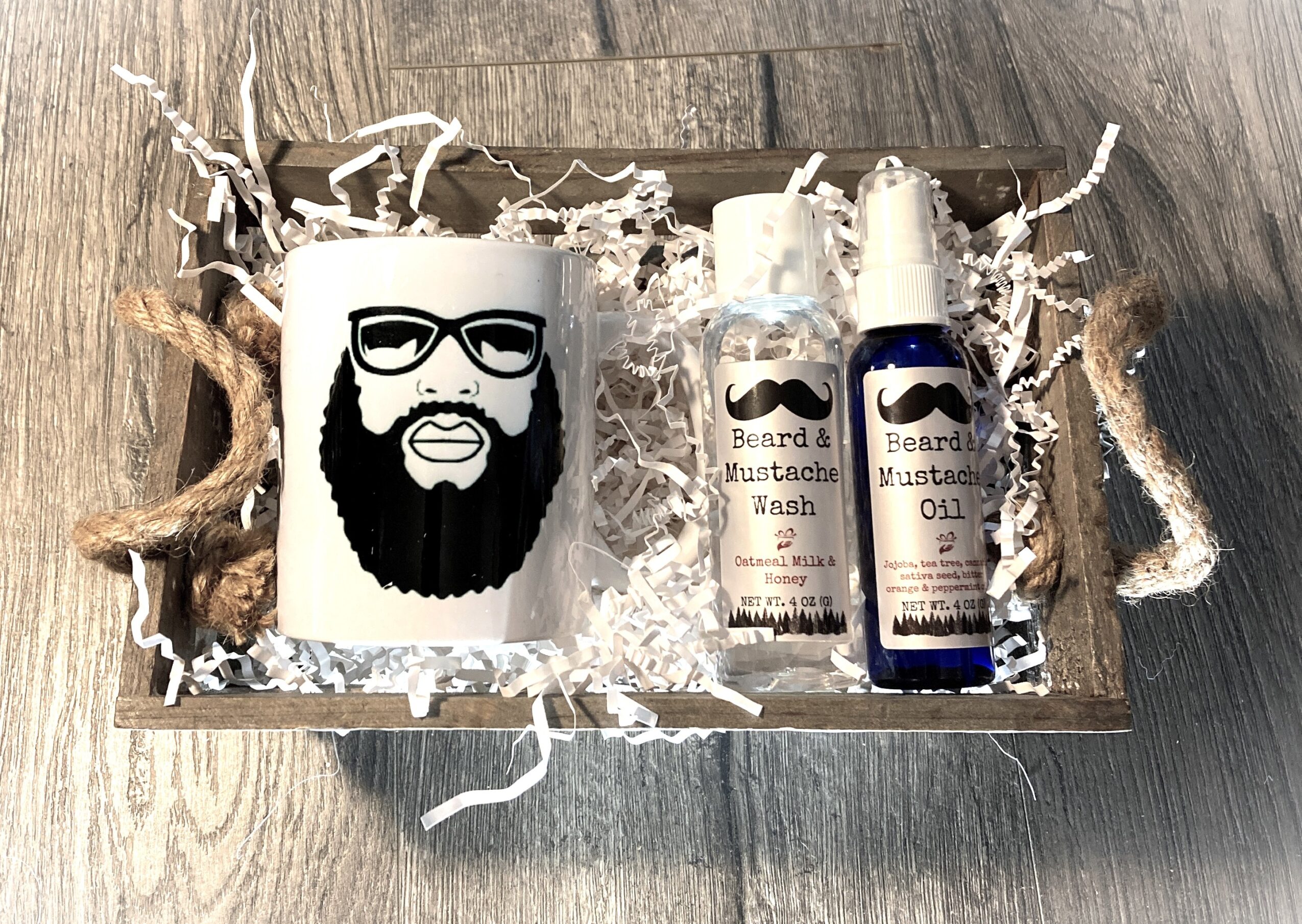 Gift box with beard-themed items, including a mug with a beard and glasses, beard wash, and beard oil, on a wooden surface.