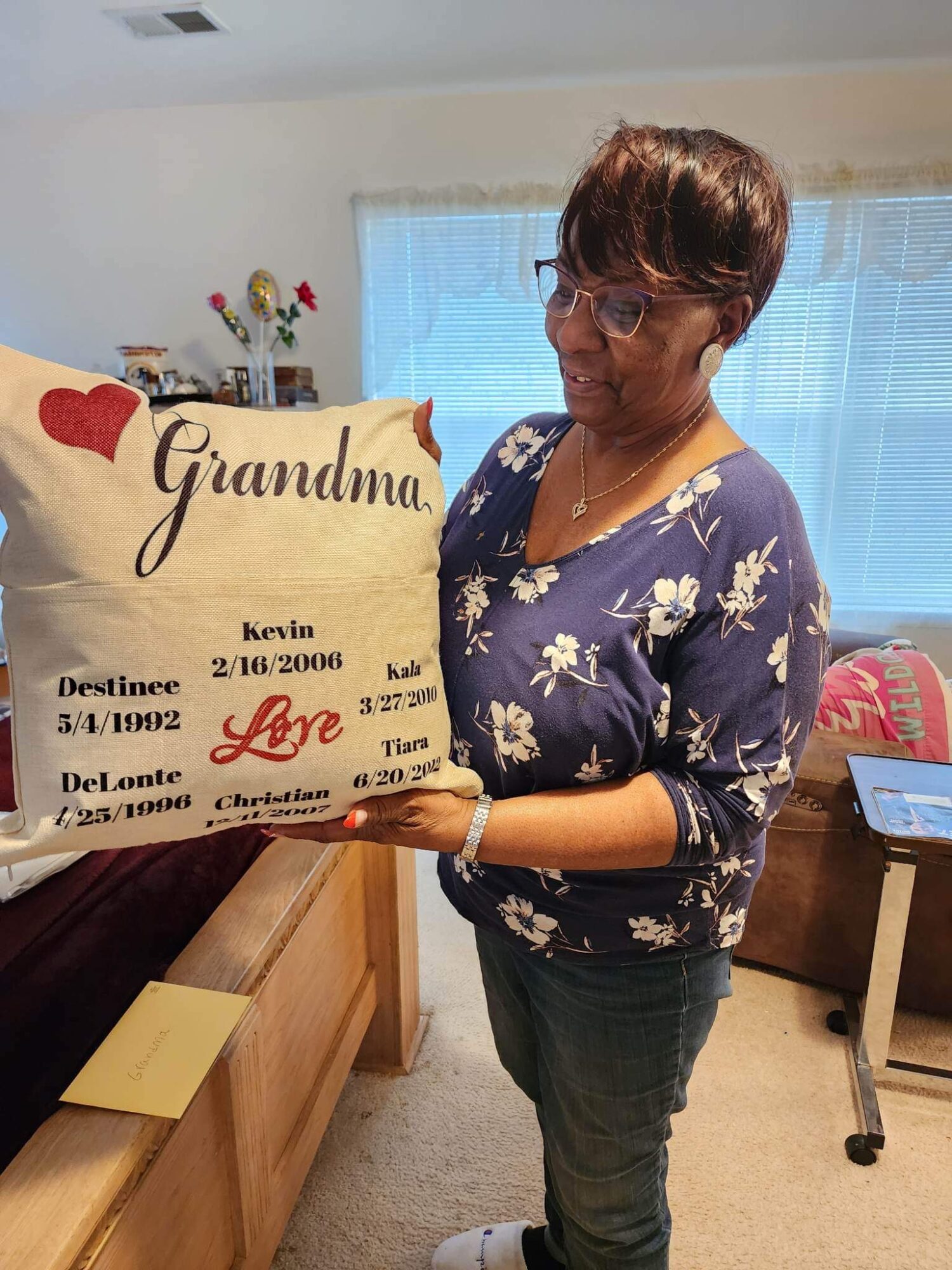 Woman with short hair and glasses holding a large decorative pillow with text and hearts, standing in a room with a window and furniture.