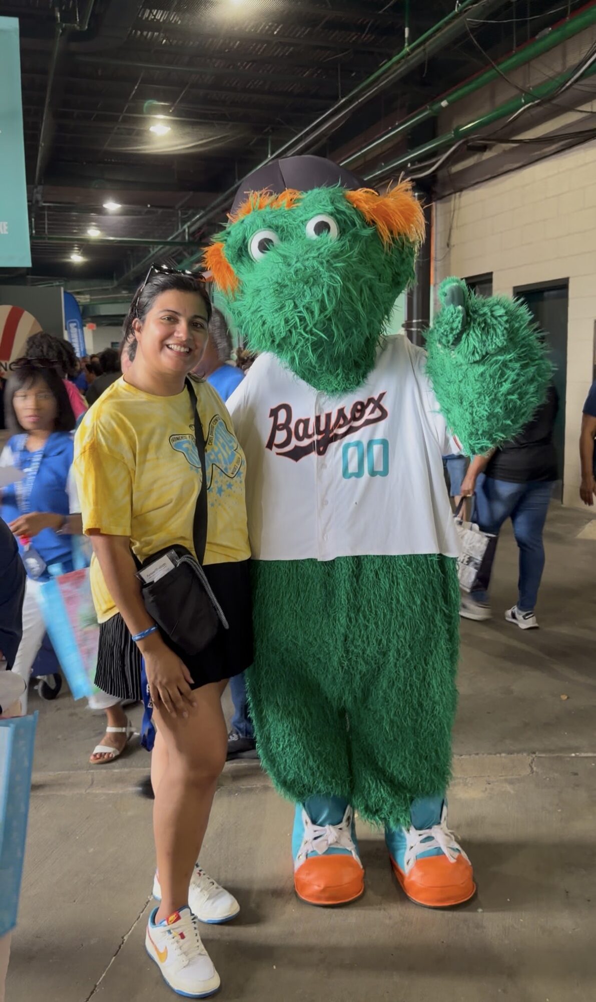 Person in yellow shirt standing next to a green mascot with orange hair, wearing a white jersey, in an indoor setting.