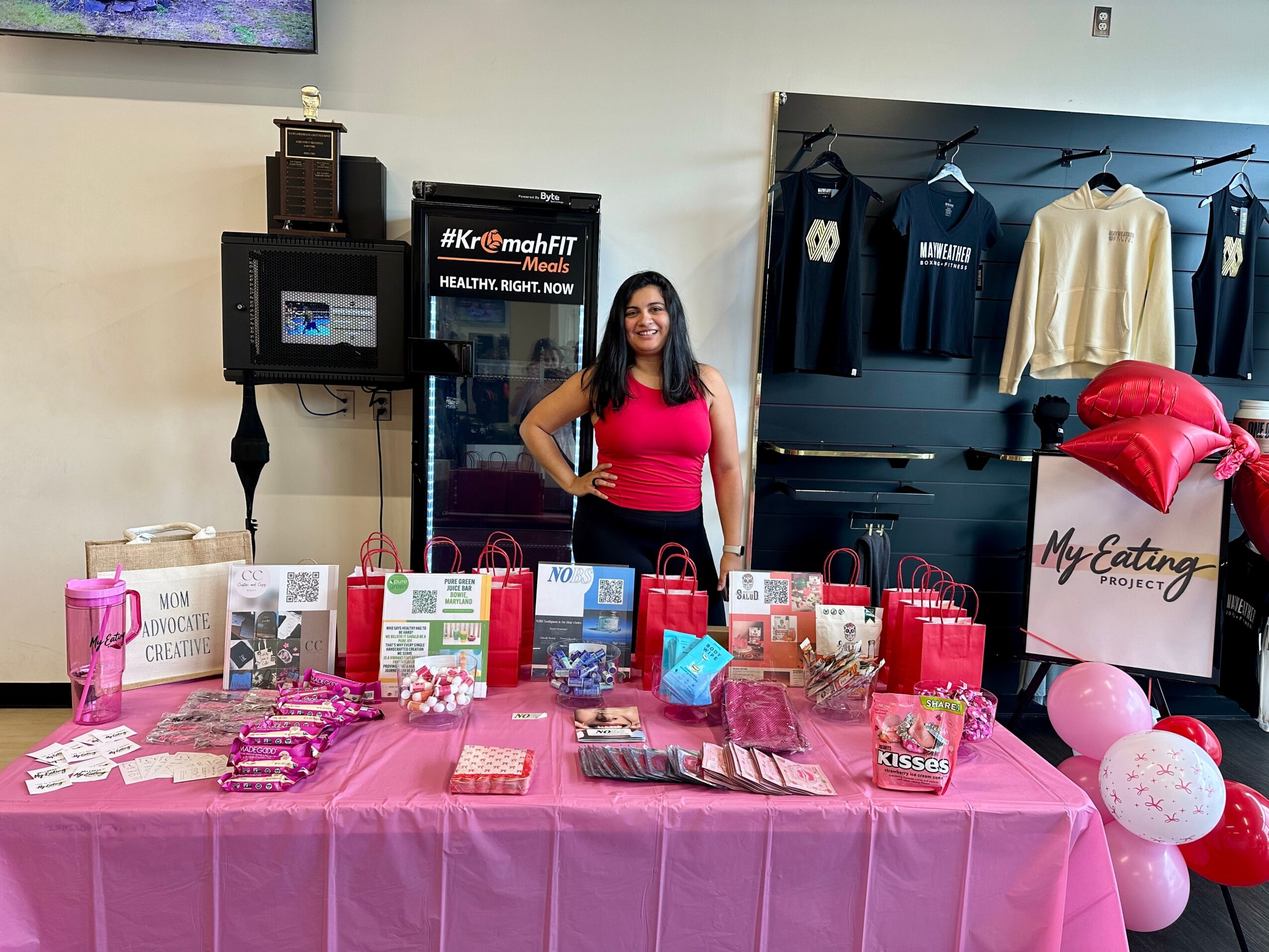 Woman standing behind a table with pink tablecloth, various gift boxes, and balloons, in a room with clothing display and equipment.