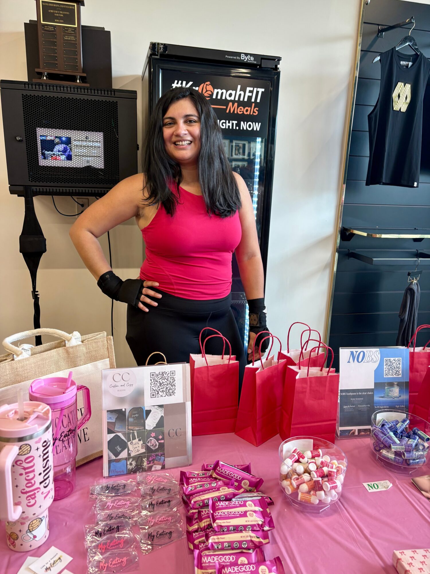 Smiling woman in pink top standing behind table with red gift bags and candy, in a store or event setting.