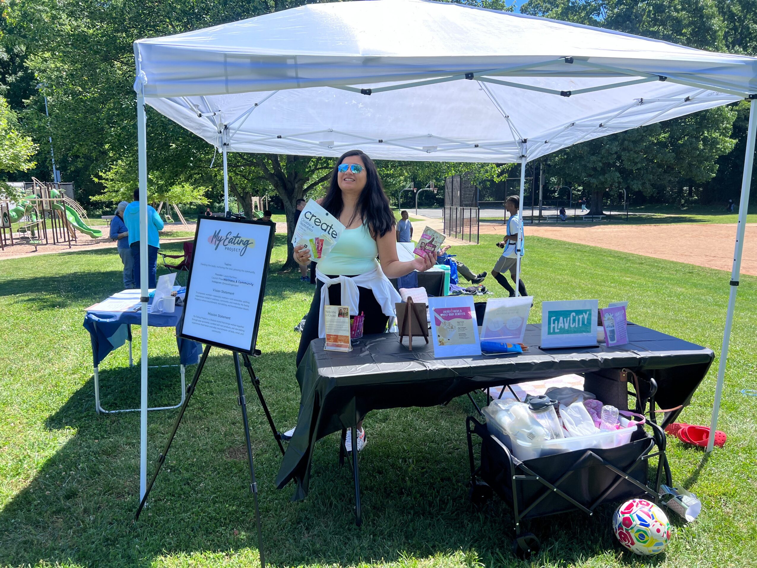 Woman standing under a white canopy at an outdoor event booth with a table displaying items, a signboard, and a grassy park background.