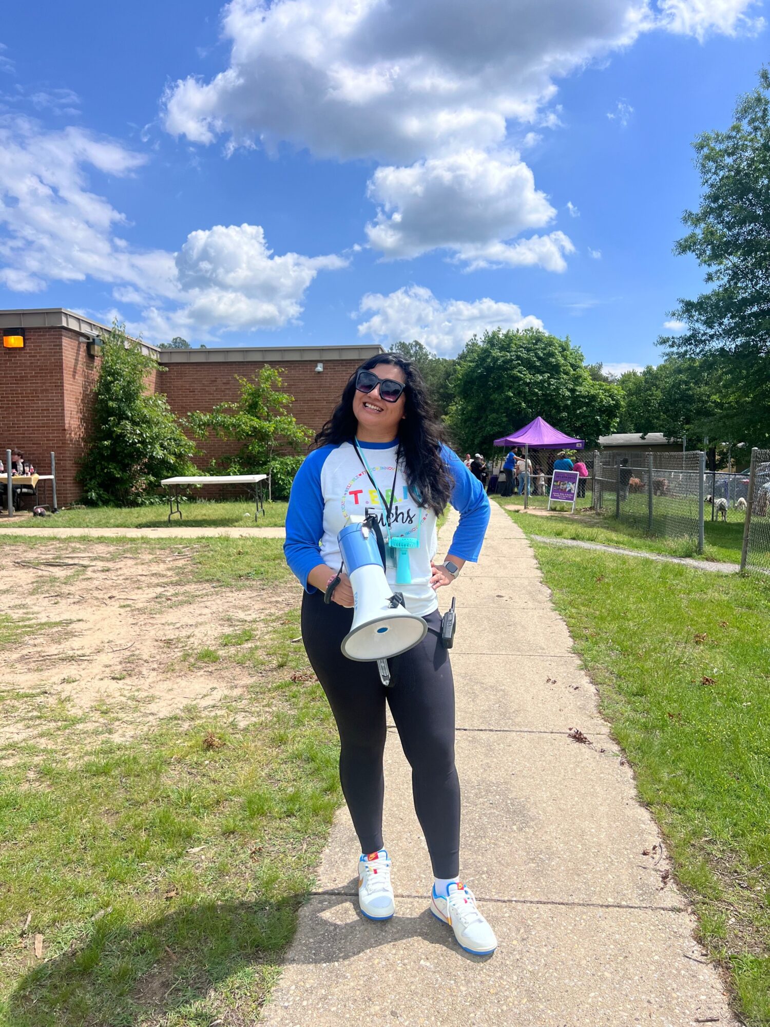 Woman standing outdoors on a sidewalk holding a white cup, with trees and a blue sky in the background.