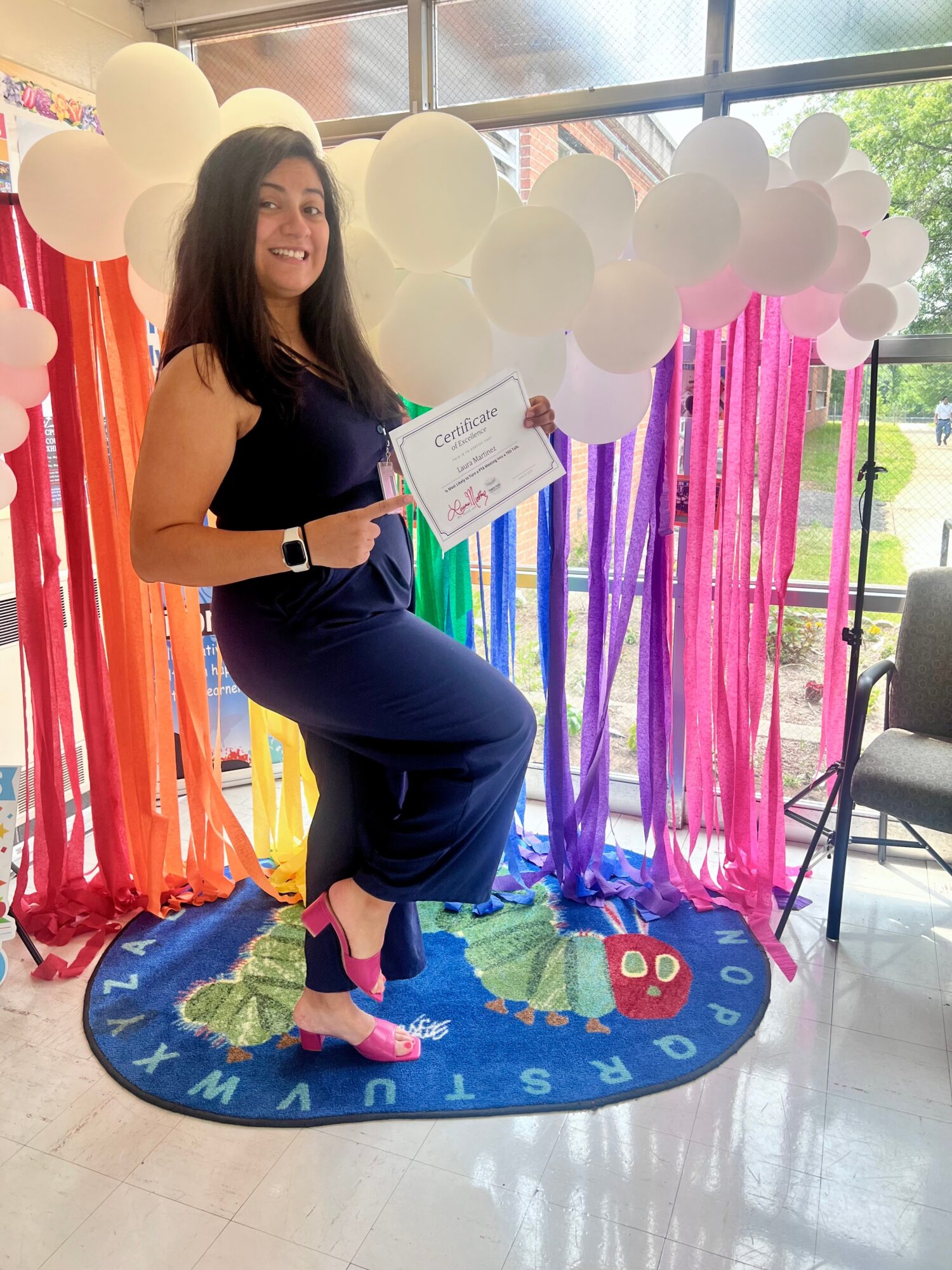 Woman in a navy dress holding a certificate, standing on a colorful rainbow-themed rug with balloons and streamers behind her.