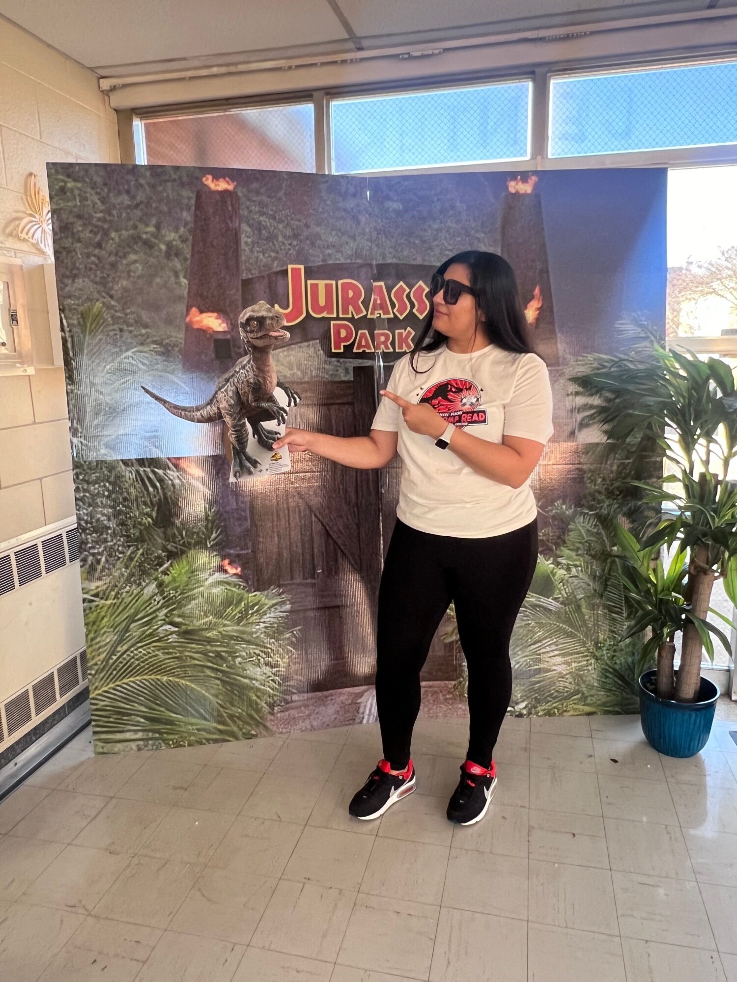 Person standing indoors in front of a Jurassic Park poster, holding a phone, with plants nearby.