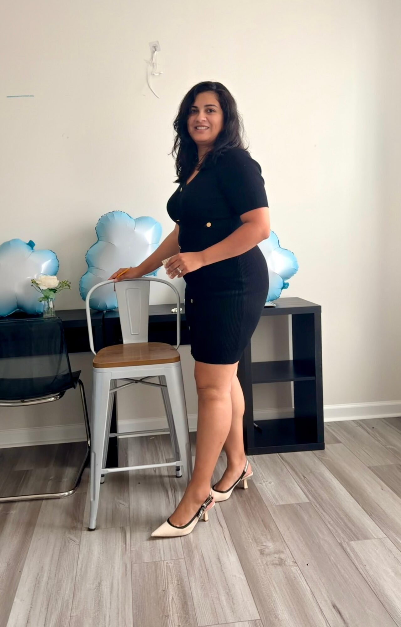 Woman in black dress and heels standing next to a chair with a glass of water, in a room with balloons and a black shelf.
