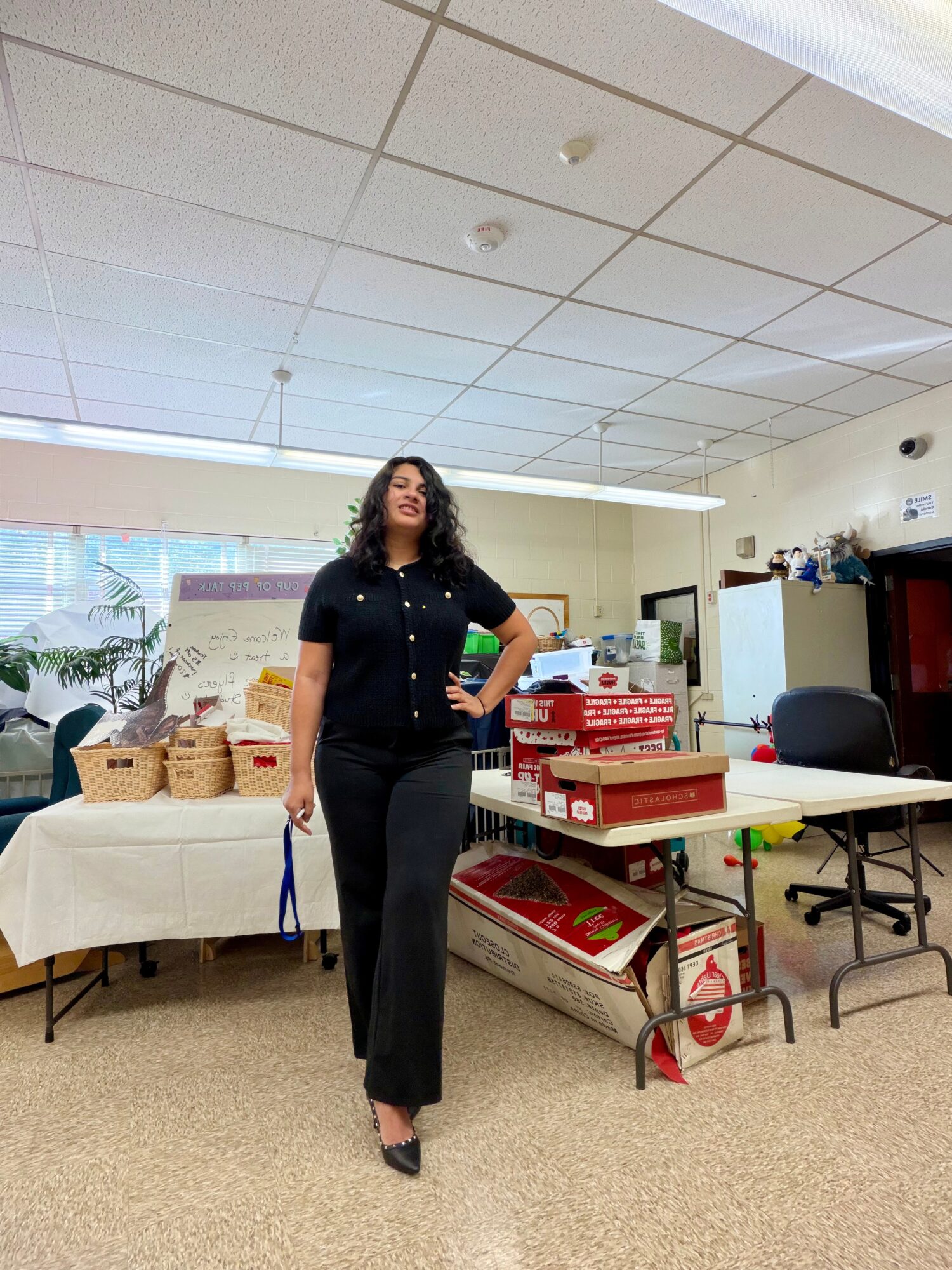 Woman standing in an office or classroom with tables, chairs, and boxes, wearing a black outfit and high heels.
