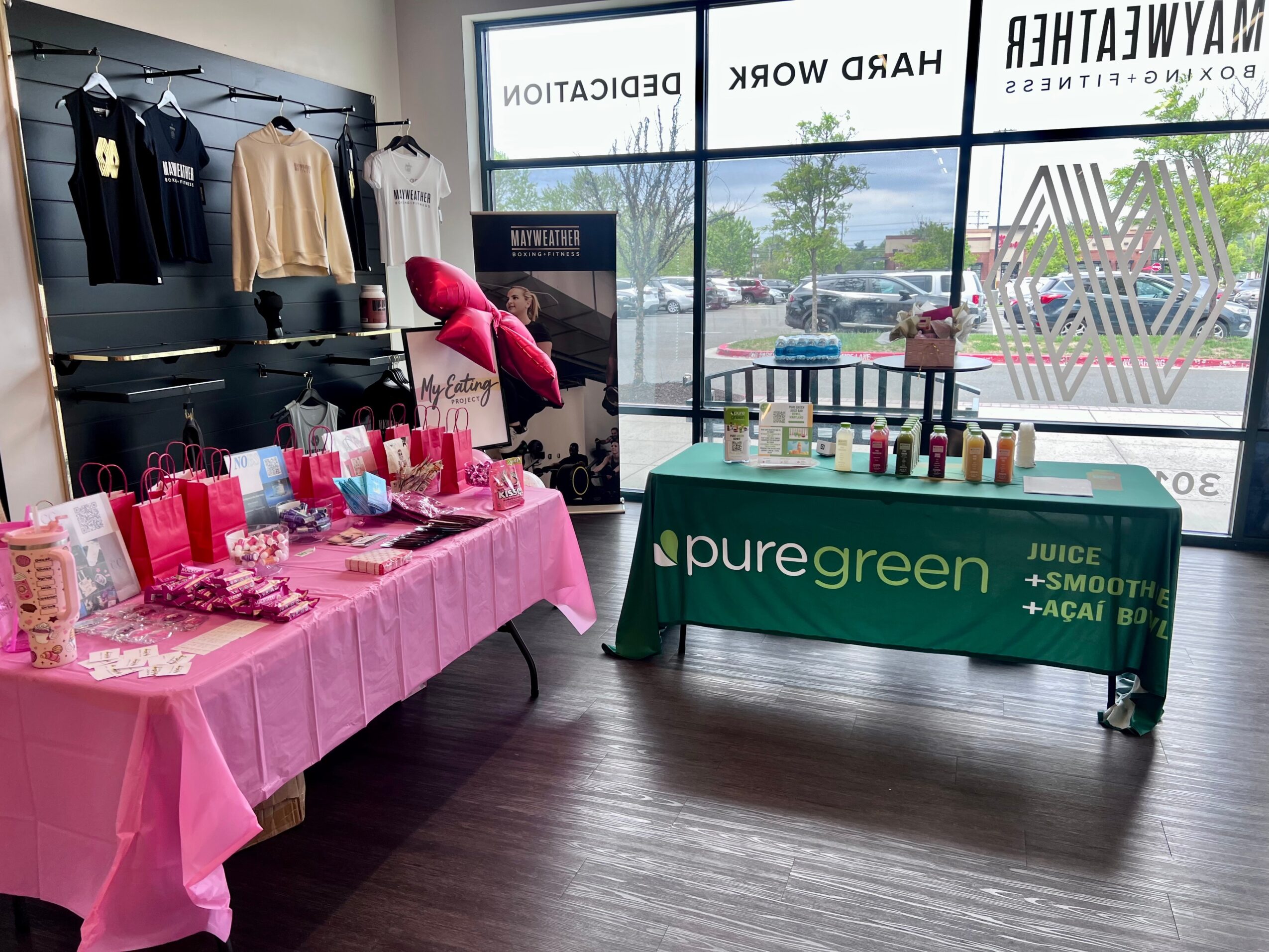 Indoor display with pink tablecloths, gift bags, and a green table with juice bottles, near large windows showing parking lot.