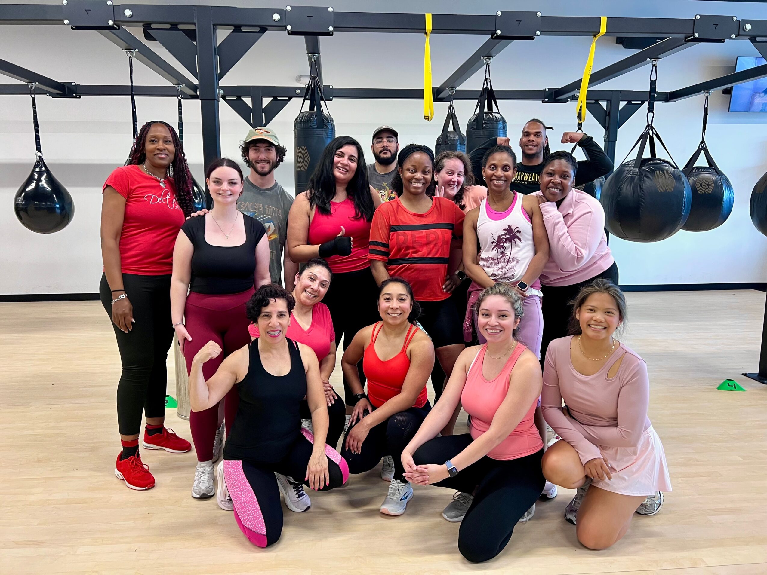 Group of people in a gym, some standing and some kneeling, smiling, with boxing bags in the background.
