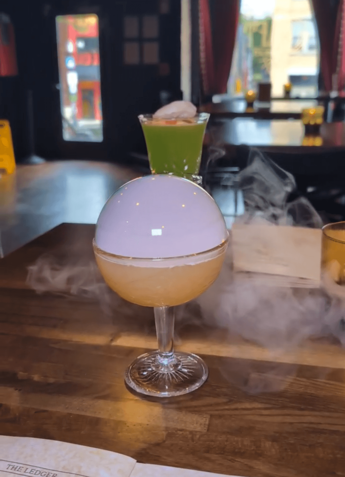 A cocktail with a frothy top and smoke effects in a glass, with a green drink in a shot glass behind it.