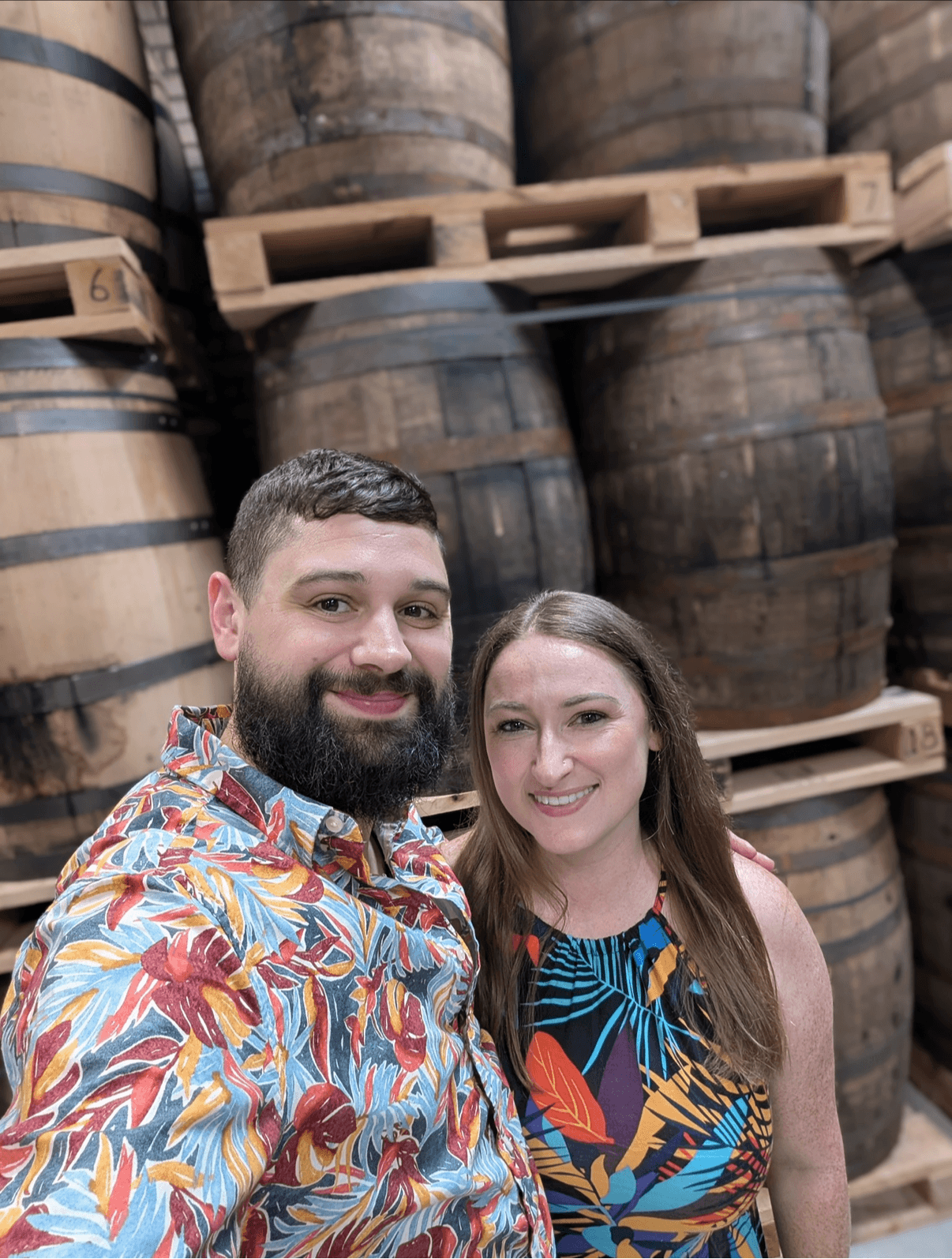 A man and woman smiling in front of stacked barrels in a warehouse, both wearing colorful patterned clothing.