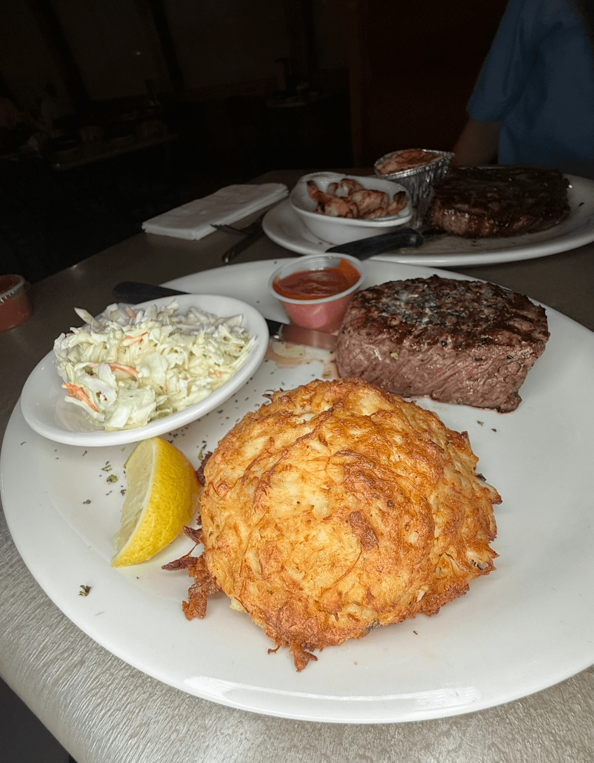 Plate with fried hash brown, lemon wedge, coleslaw, and small container of ketchup, on a table with other dishes in background.