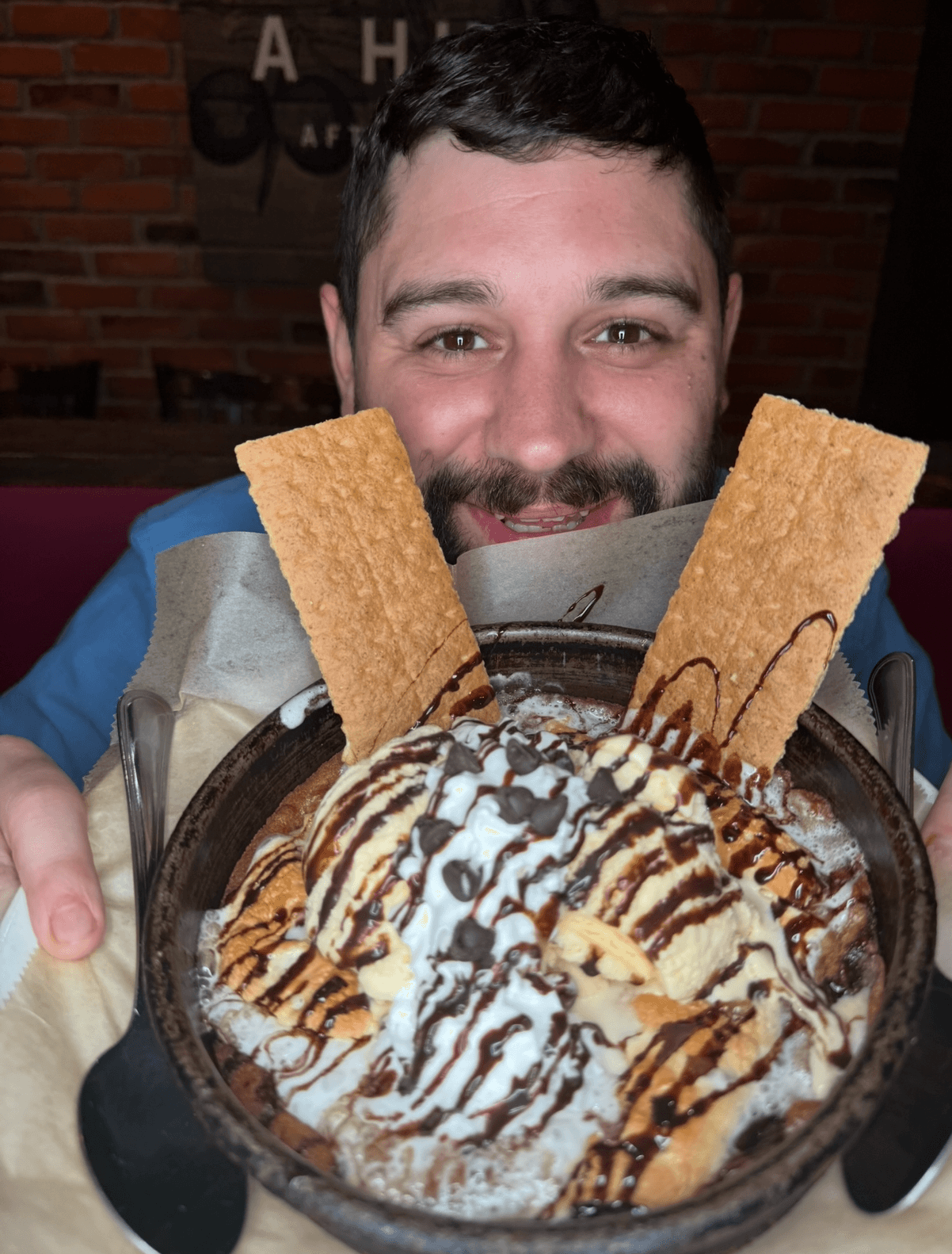 Person smiling with a dessert topped with ice cream, chocolate drizzle, and crispy wafer sticks.