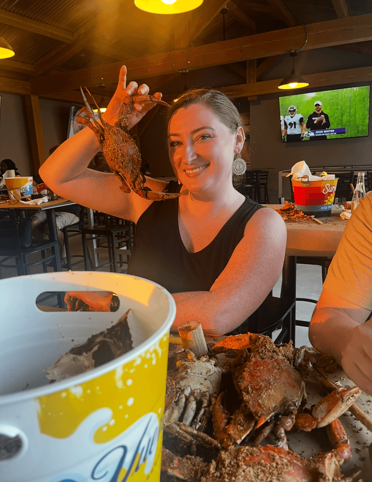 Woman smiling and holding a large cooked lobster in a restaurant setting with food and drinks on the table.