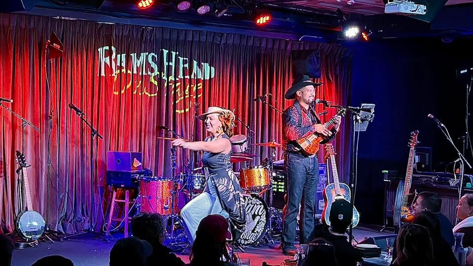 Two musicians perform on stage with red curtains and a sign reading 'Films Head' behind them.