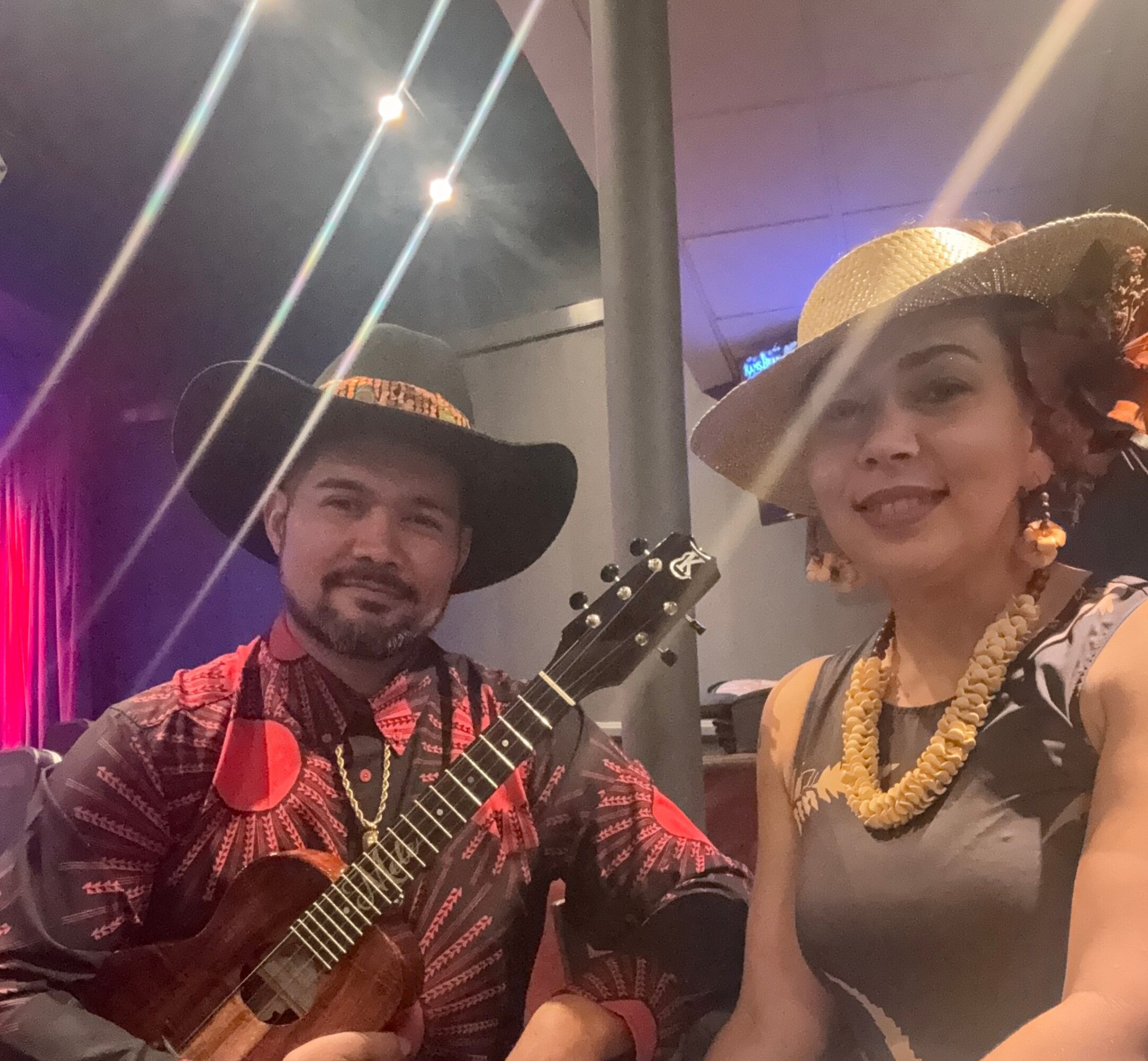 Two people dressed in traditional attire, one holding a guitar, smiling indoors with colorful lighting.