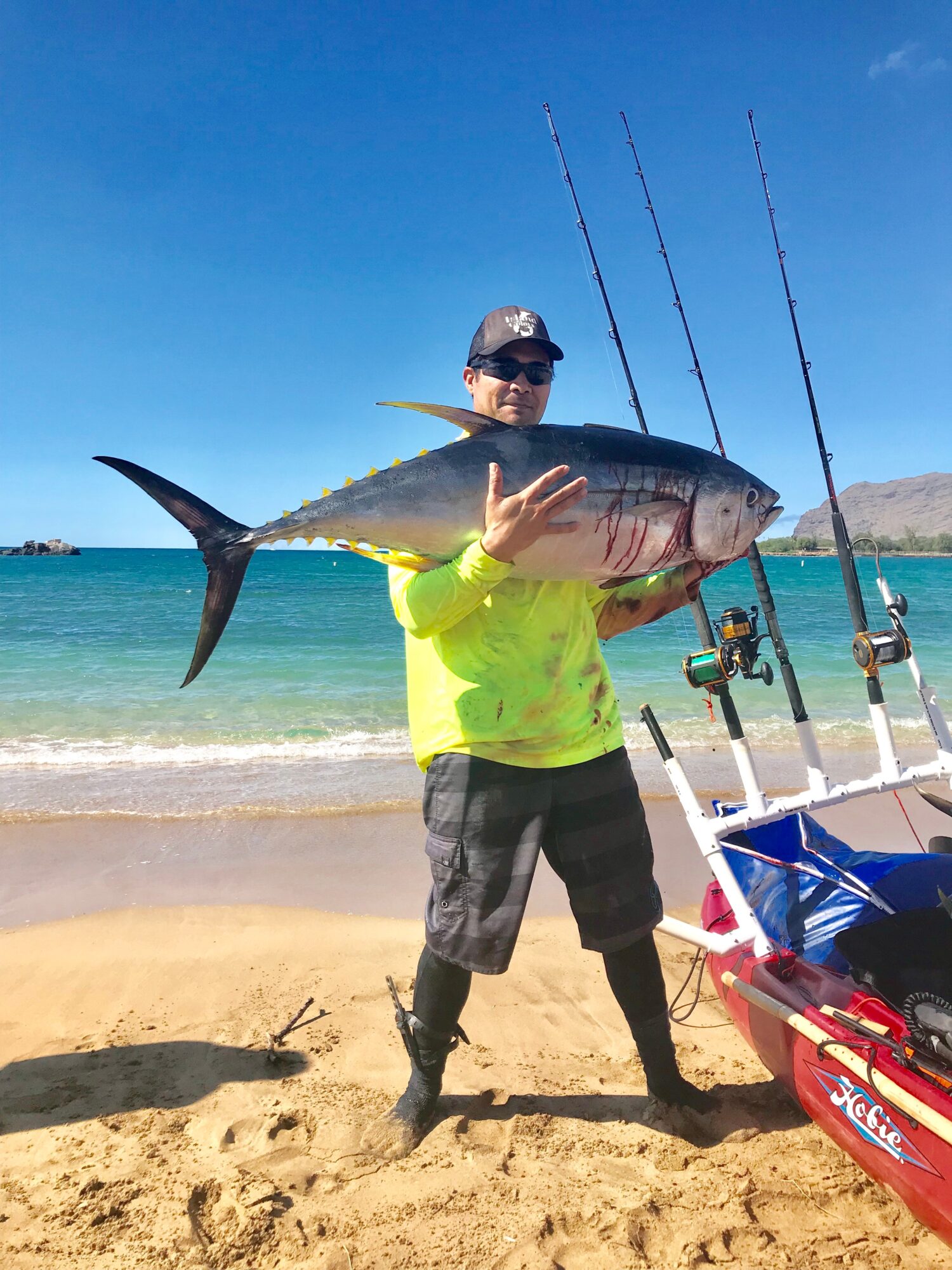 Person holding a large fish on a sandy beach with the ocean in the background, two fishing rods, and a blue and red fishing cart.