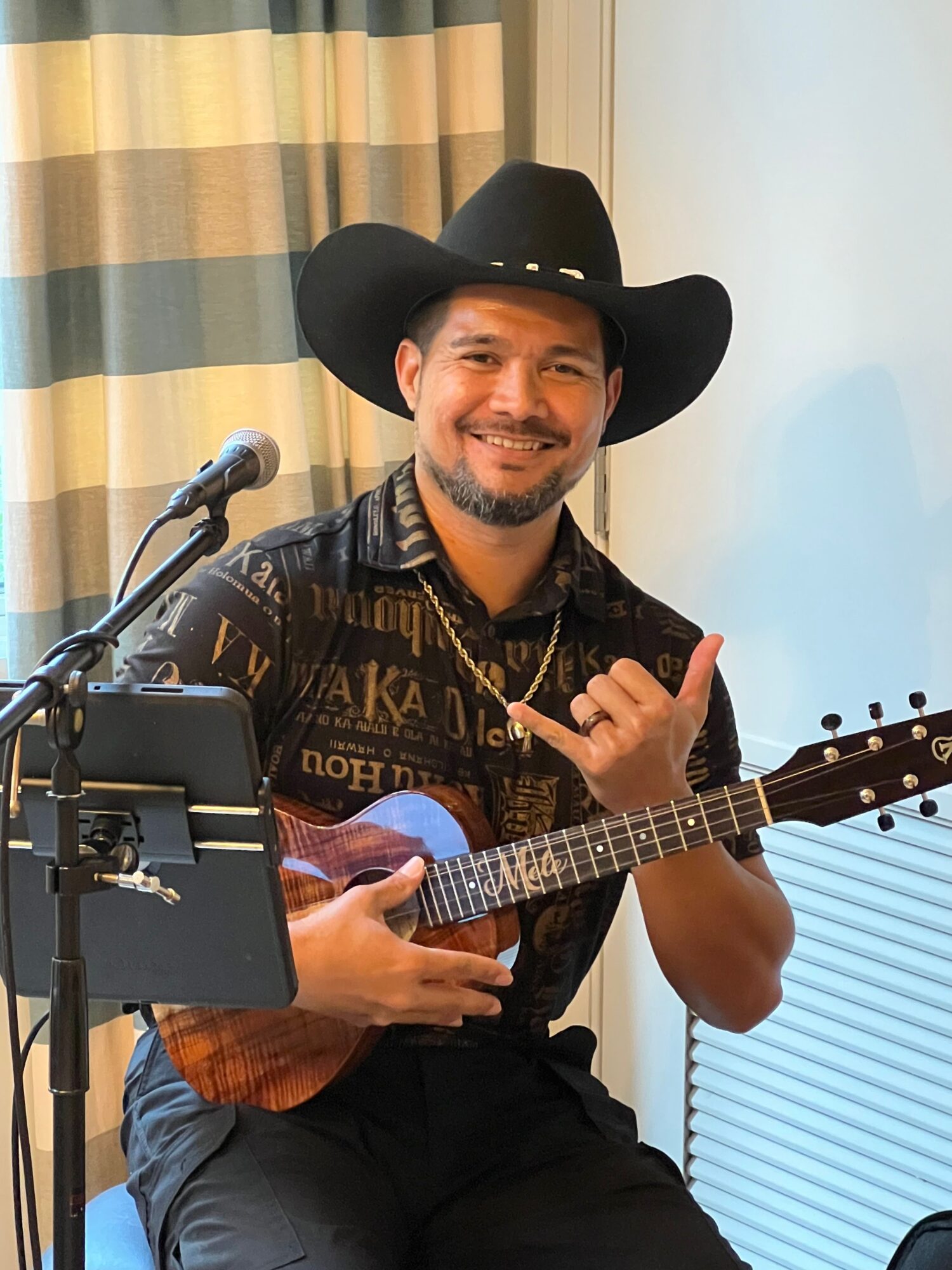 Man wearing a cowboy hat playing an acoustic guitar, sitting near a window with blinds, smiling at the camera.