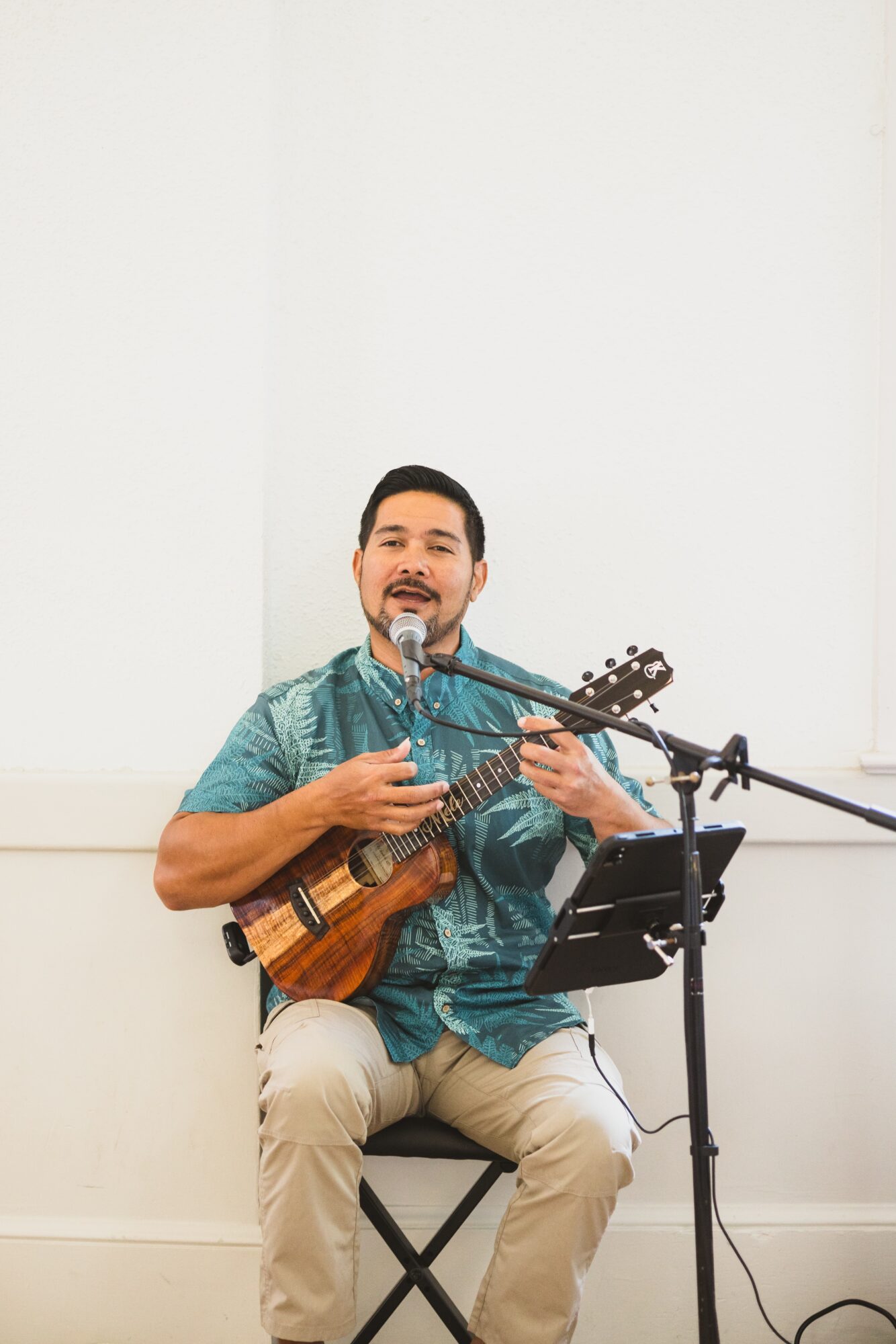 Man playing ukulele and singing into a microphone, seated with music stand nearby, against a plain white wall.