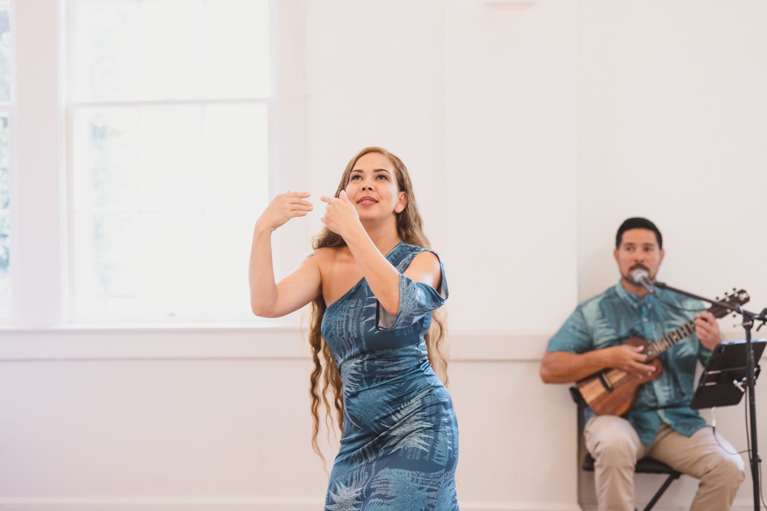 Woman with long hair in a blue dress dancing, man playing guitar in background, bright room with large window