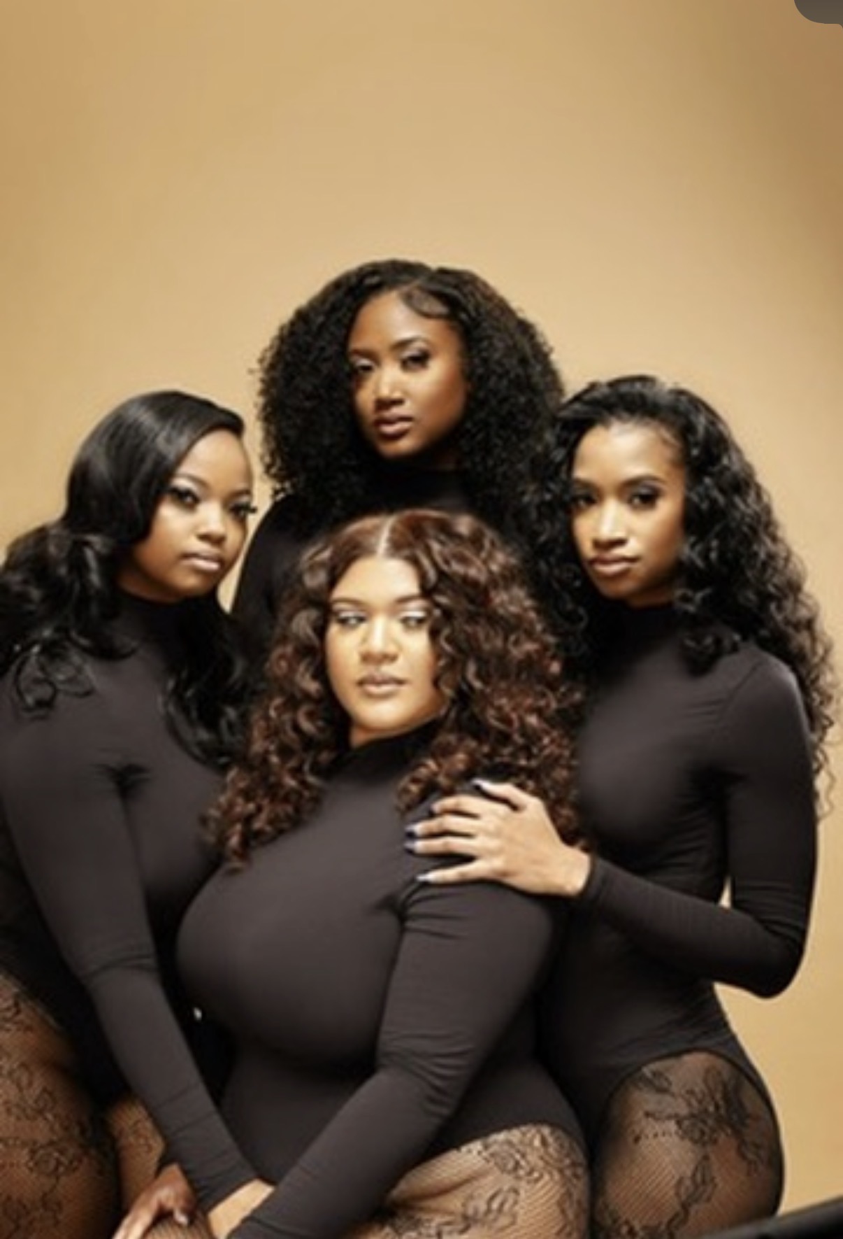 Four women with curly and straight hair pose together against a beige background, wearing black outfits. One woman rests her hand on another's shoulder.