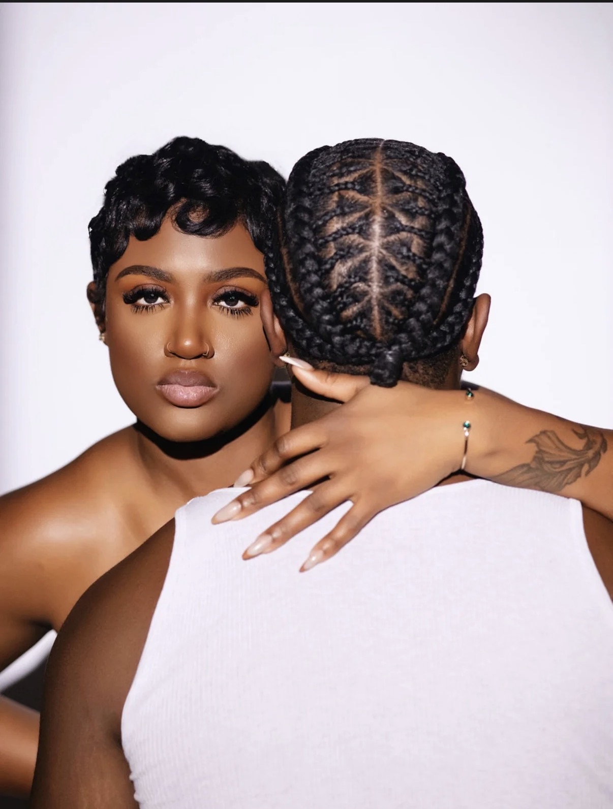 Two women with braided hairstyles face each other, one with visible face and the other with back view, against a plain background.