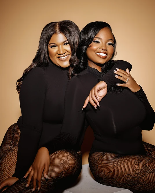 Two women with dark hair and black outfits sitting close together, smiling, against a beige background.