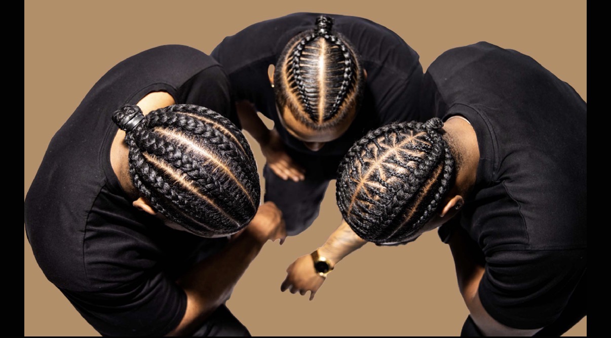 Three people with braided hairstyles in black clothing form a circle, viewed from above, against a brown background.