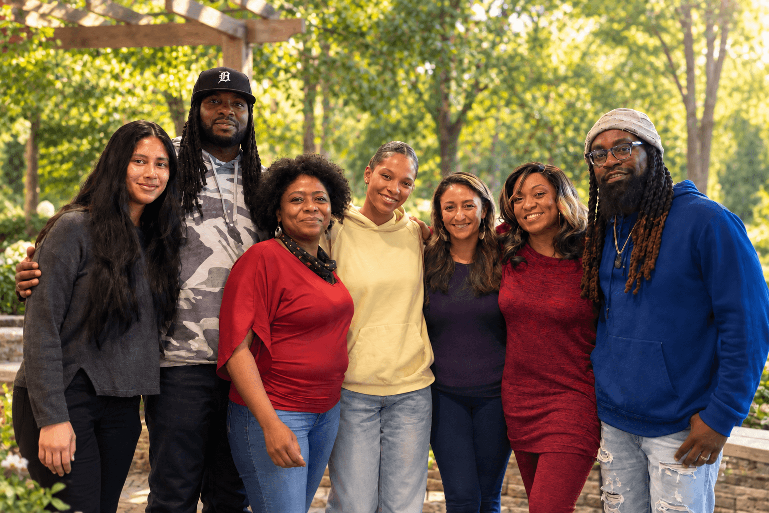 Group of seven diverse people standing outdoors in a park, smiling, with trees and sunlight in the background.