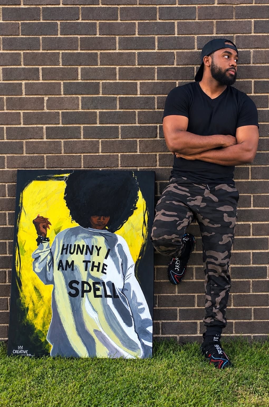 Man with arms crossed leaning against a brick wall next to a colorful painting with text, wearing a black shirt and camouflage pants.