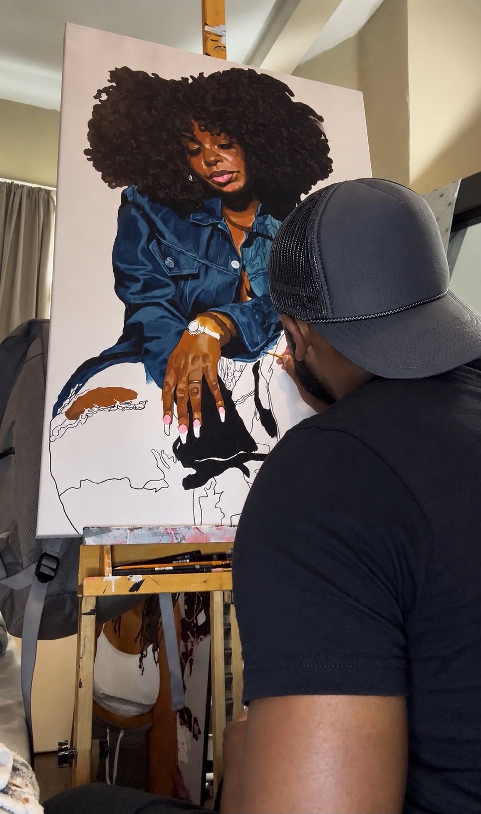 Person with curly hair painting a portrait of a woman with curly hair wearing a blue jacket, on an easel.