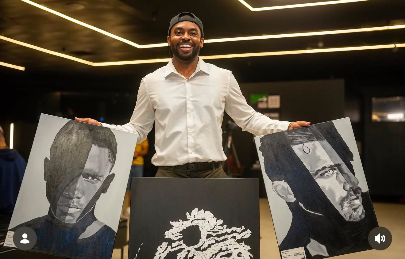Man smiling standing behind artwork displays in an indoor gallery or event space.