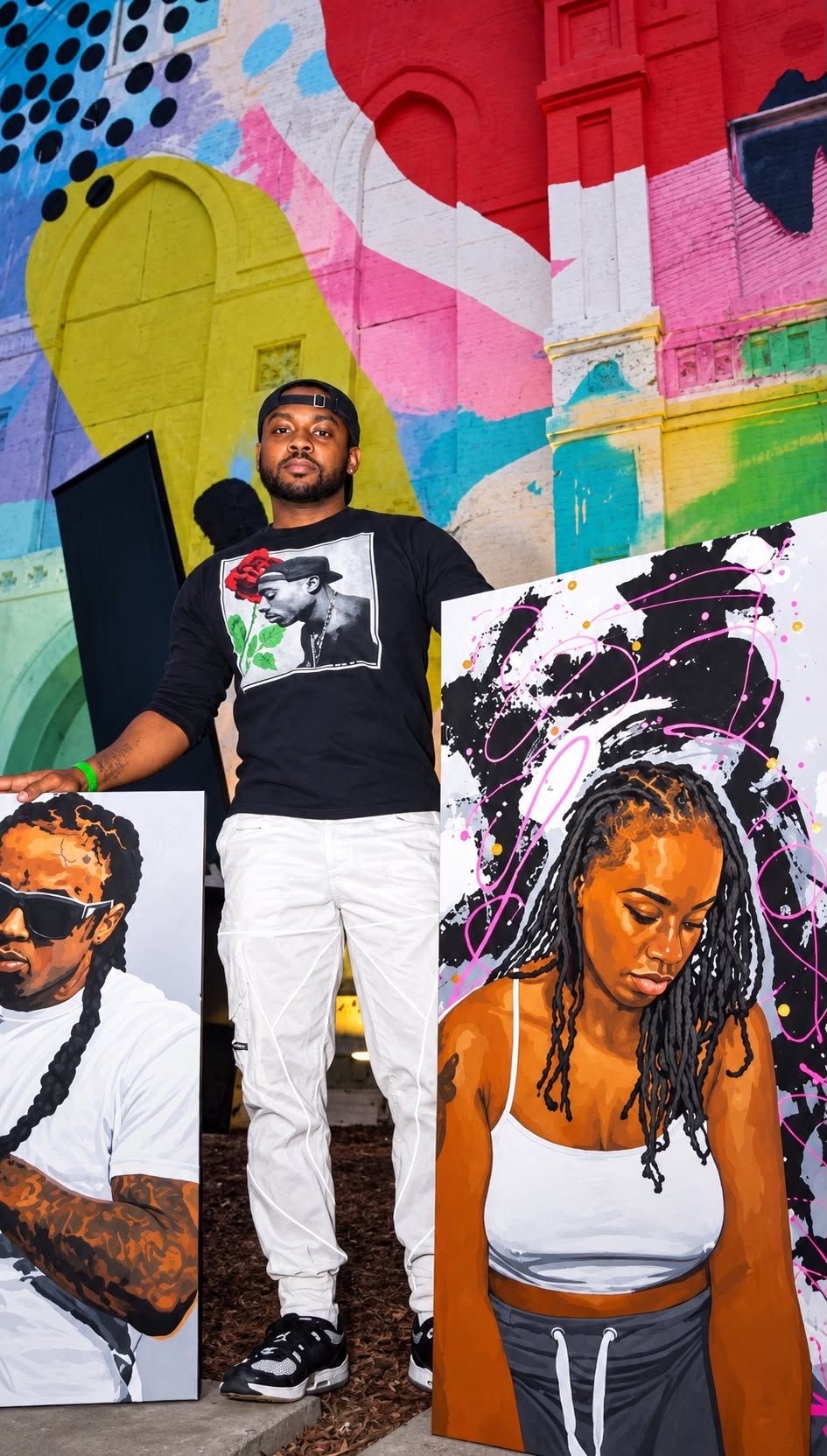 Man standing in front of colorful graffiti wall, with two large portrait paintings of women on either side.