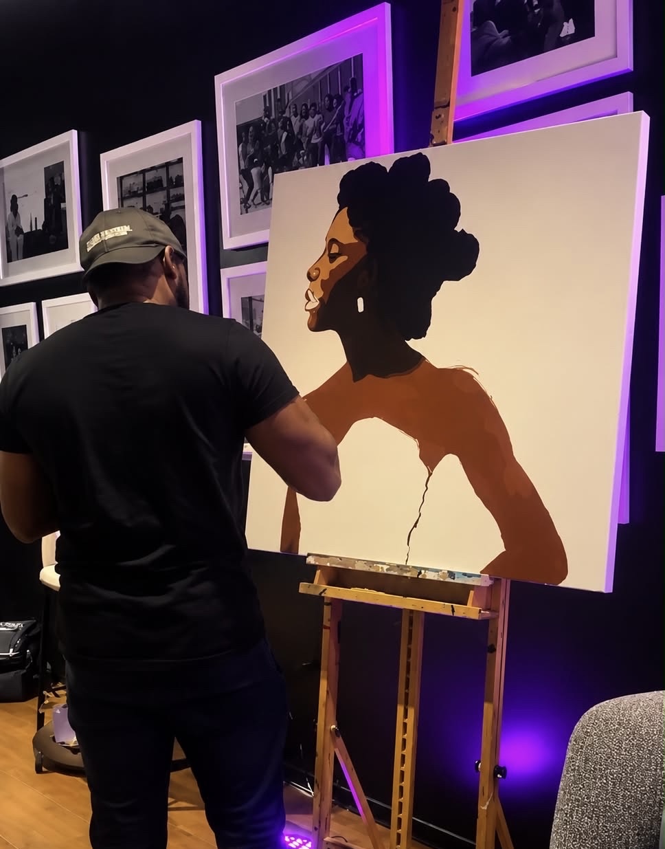 Artist painting a portrait of a woman with an elaborate hairstyle on a large canvas, in a gallery with framed photos and purple lighting.